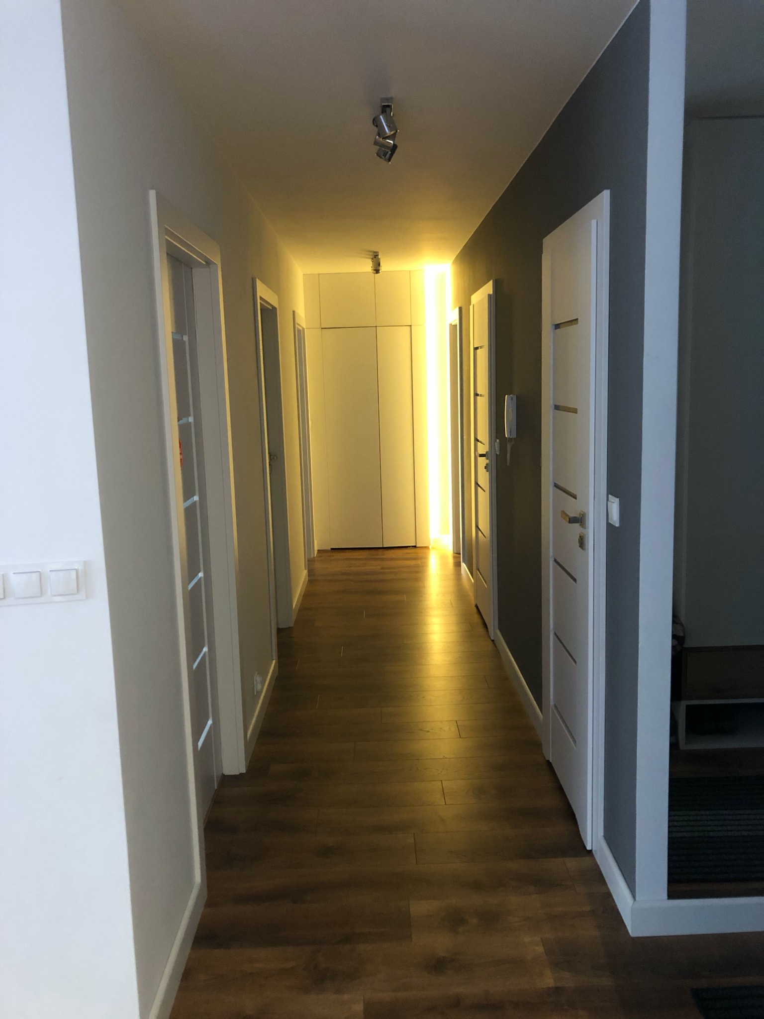 Długi korytarz z drewnianą podłogą, białymi drzwiami i oświetleniem LED w zabudowie meblowej na końcu, ściany pomalowane na biało i szaro.