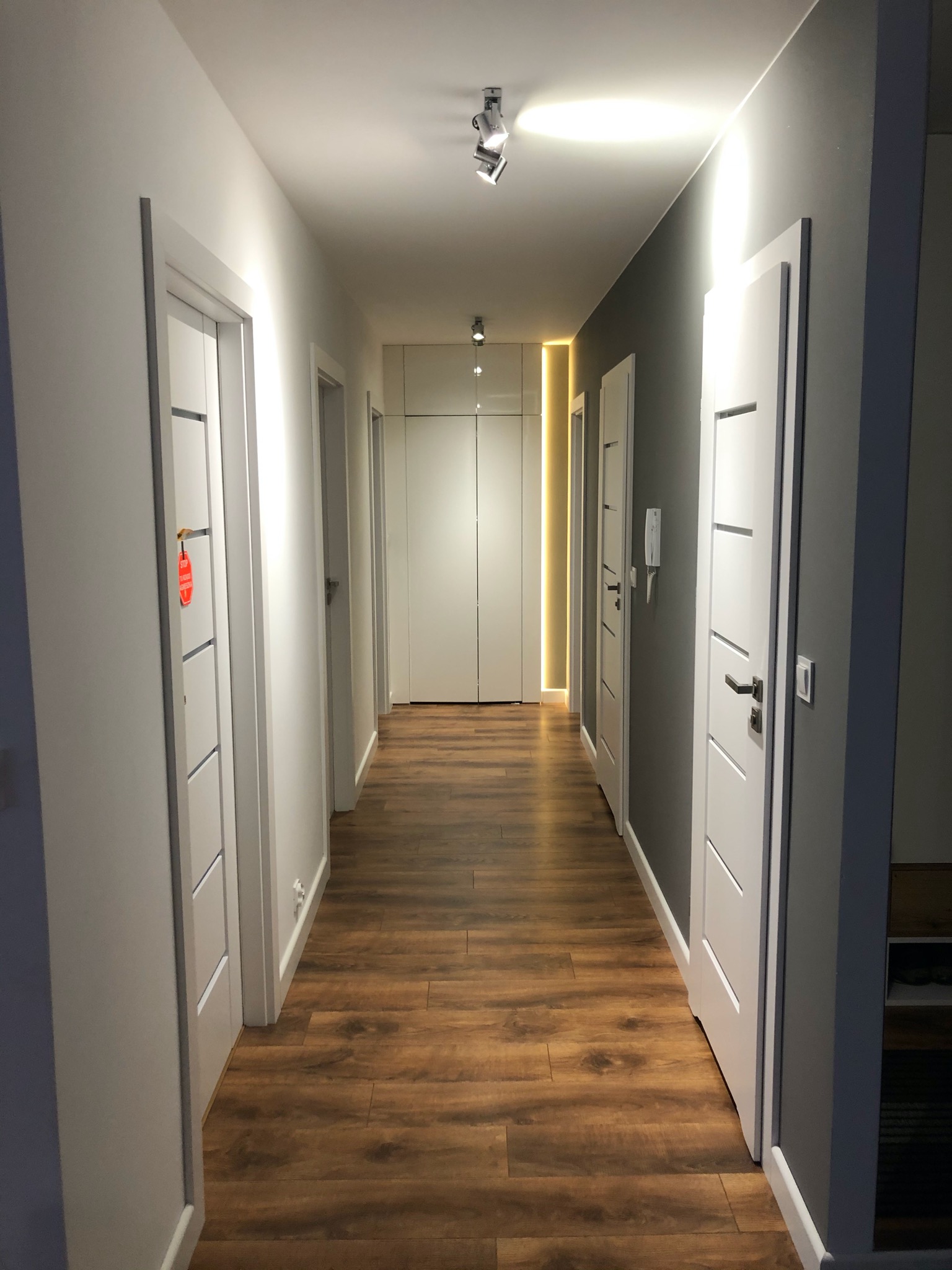 Długi korytarz z drewnopodobną podłogą, białymi drzwiami z poziomymi frezami, szarą ścianą po prawej stronie i oświetleniem LED wzdłuż ściany.