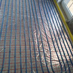 Niebieskie rury ogrzewania podłogowego rozłożone na folii z siatką w pomieszczeniu, widoczne okno w tle.