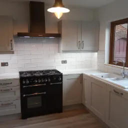 Jasna kuchnia z białymi płytkami, czarną kuchenką i drewnianą ramą okienną z widokiem na ceglany budynek.