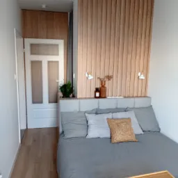 Nowoczesna sypialnia w neutralnych kolorach z drewnianą ścianą dekoracyjną, łóżkiem z szarą pościelą i śniadaniem na tacy.