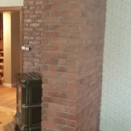 Wykończony fragment wnętrza z ceglaną kolumną i obudową kominka, drewnianą podłogą w parkiet oraz dekoracyjną listwą przysufitową.
