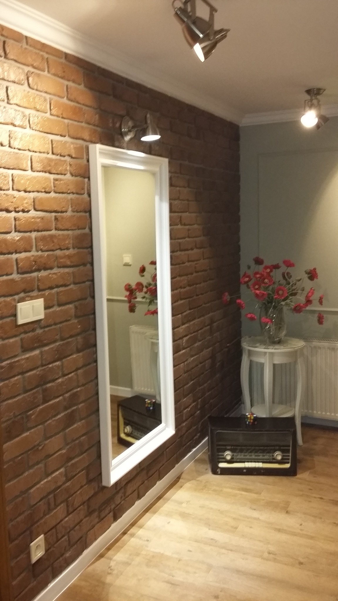 Wnętrze pokoju z ceglaną ścianą, dużym lustrem w białej ramie odbijającym fragment pokoju, drewnianą podłogą i staromodnym radiem na podłodze.