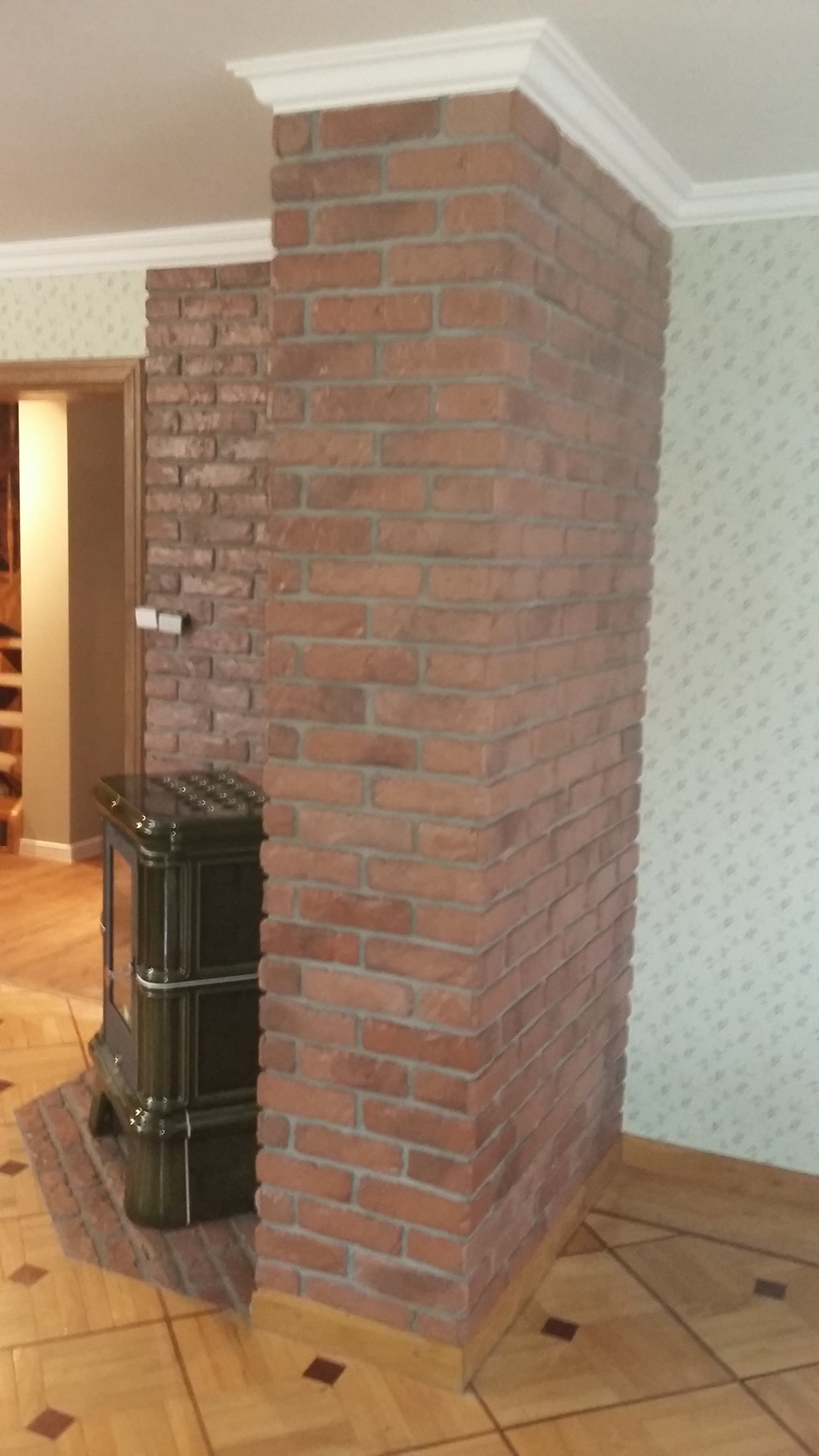 Wykończony fragment wnętrza z ceglaną kolumną i obudową kominka, drewnianą podłogą w parkiet oraz dekoracyjną listwą przysufitową.