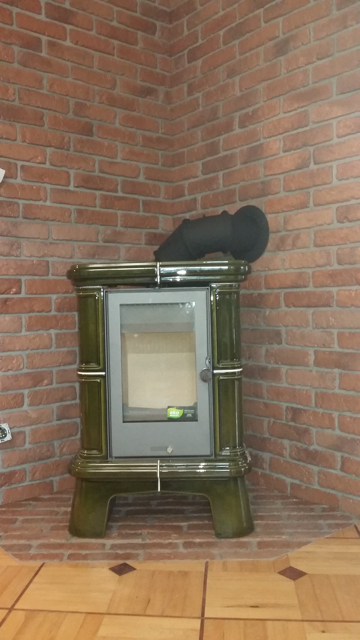 Zielony piec kaflowy z czarną rurą odprowadzającą dym, ustawiony w rogu pomieszczenia z ceglaną ścianą i drewnianą podłogą z ciemnymi akcentami.
