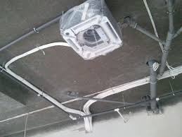 Kasetonowa jednostka klimatyzacji w trakcie montażu, zabezpieczona folią ochronną, widoczne rury instalacyjne na szarym suficie.