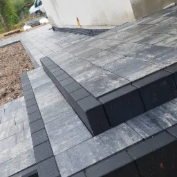 Schody i chodnik z szarej kostki brukowej z czarnym obrzeżem, prowadzące do budynku z jasną elewacją i widocznym oknem, częściowo ułożone na piasku.