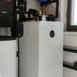 Instalacja z białą jednostką marki Bosch, czerwonym i niebieskim zbiornikiem ciśnieniowym oraz czarnymi rurami w pomieszczeniu technicznym w Otwocku.