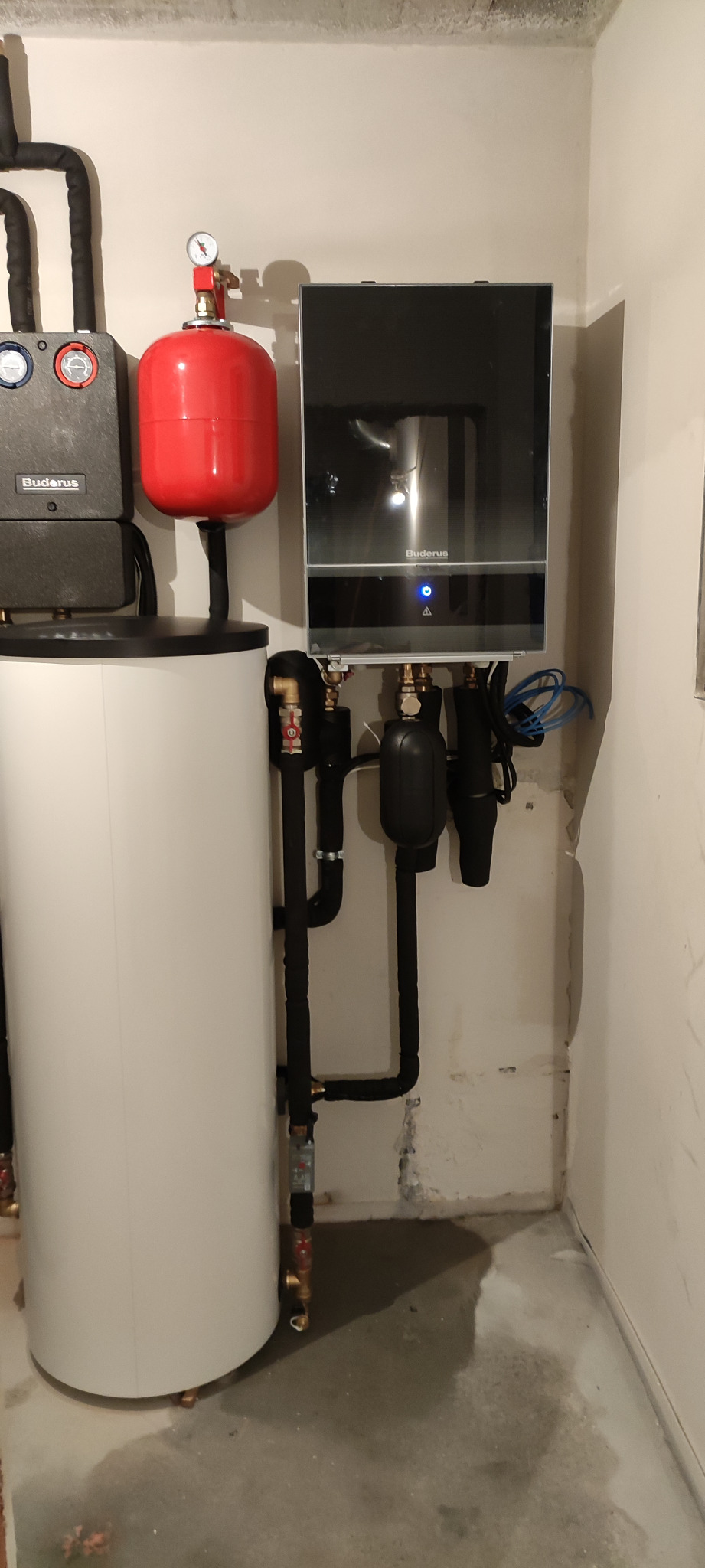 Kompletna instalacja grzewcza Buderus z zasobnikiem ciepłej wody, czerwonym zbiornikiem wyrównawczym i czarnymi rurami w izolacji, zamontowana przy ścianie w pomieszczeniu technicznym.