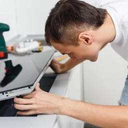 Mężczyzna w białej koszulce instaluje płytę indukcyjną w białej zabudowie kuchennej, w tle wiertarka akumulatorowa.