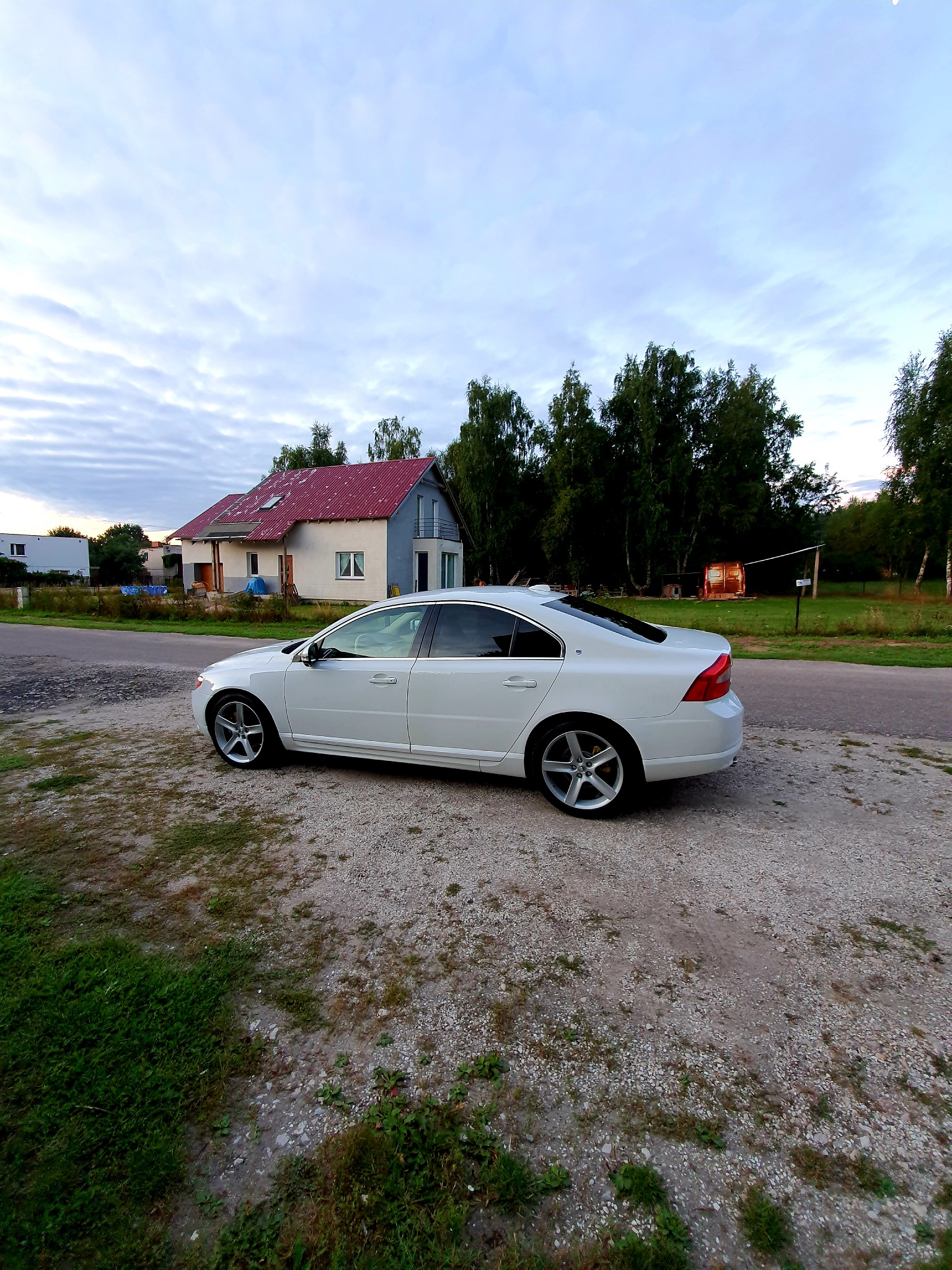Biały samochód osobowy Volvo S80 zaparkowany na żwirowym podłożu, z domem jednorodzinnym z czerwonym dachem i zielonymi drzewami w tle, pochmurne niebo.
