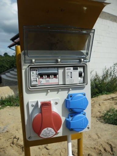 Zewnętrzna skrzynka elektryczna z bezpiecznikami i gniazdami siłowymi w kolorze czerwonym i niebieskim, zamontowana na żółtym słupku, z otwartą osłoną zabezpieczającą.