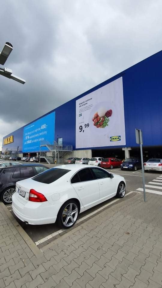 Biały samochód marki Volvo zaparkowany na parkingu przed budynkiem IKEA z dużym plakatem reklamowym na ścianie.