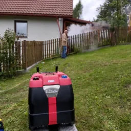 Czarno-czerwona parownica Optima Steamer na metalowych najazdach, w tle mężczyzna w jasnej kurtce czyszczący parą drewniany płot na tle domu z czerwonym dachem.