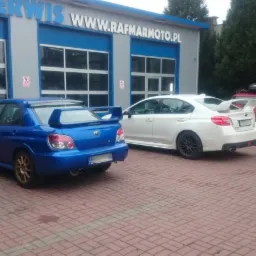 Dwa samochody Subaru, niebieski Impreza i biały WRX, zaparkowane przed warsztatem samochodowym z niebieskimi bramami i szyldem 'AUTOSERWIS WWW.RAFMARMOTO.PL'.