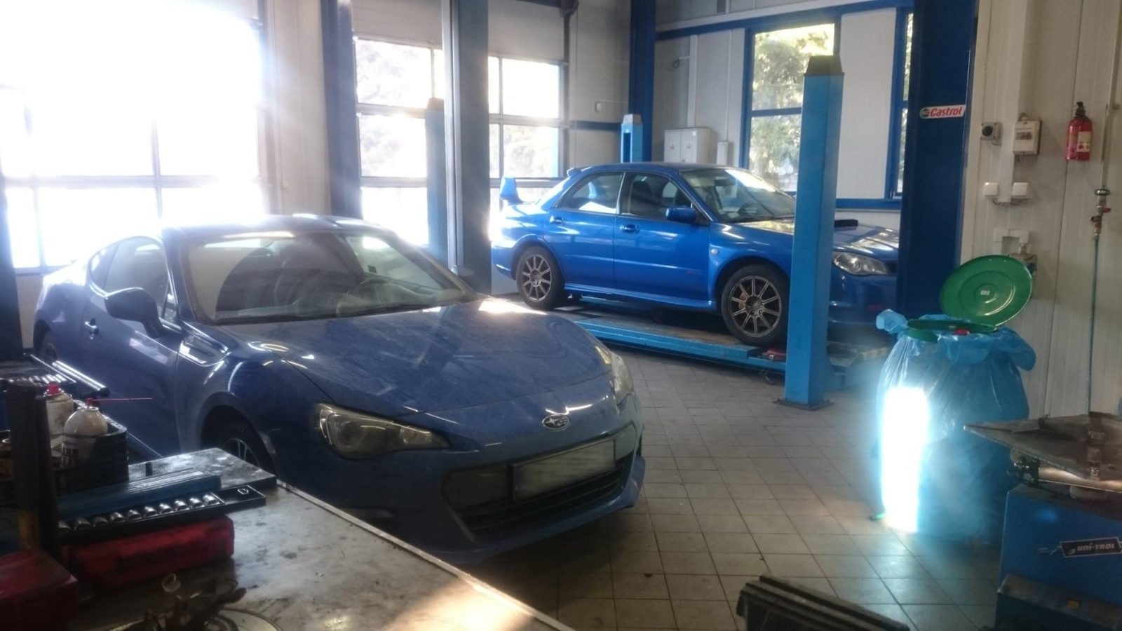 Dwa niebieskie samochody Subaru, jeden BRZ i jeden Impreza WRX, w warsztacie samochodowym, BRZ na pierwszym planie, WRX na podnośniku, narzędzia i pojemnik na śmieci w tle.