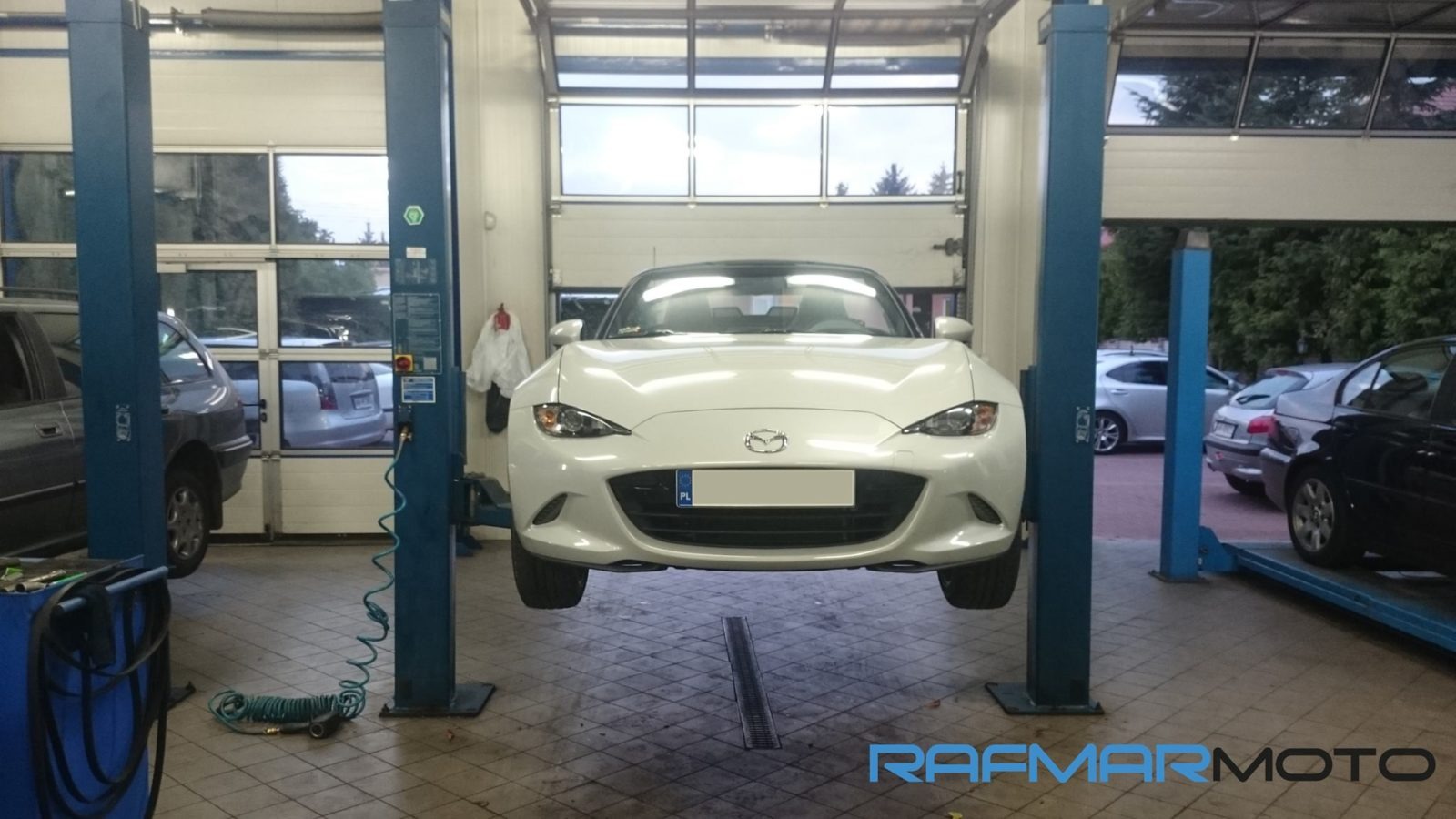 Biała Mazda MX-5 na podnośniku dwukolumnowym w warsztacie samochodowym, widok z przodu, z widocznym kanałem rewizyjnym w podłodze i innymi samochodami w tle.
