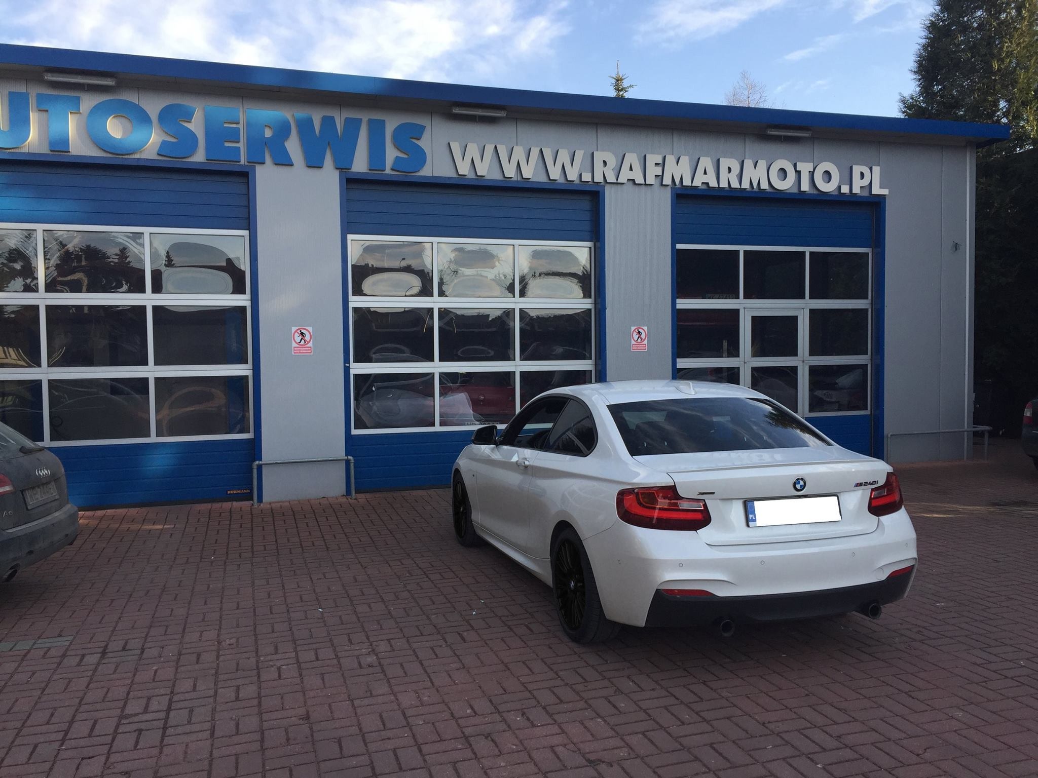 Białe BMW M240i stoi przed budynkiem warsztatu samochodowego z napisem AUTOSERWIS i adresem strony internetowej, widoczne odbicia innych aut w szybach bram wjazdowych