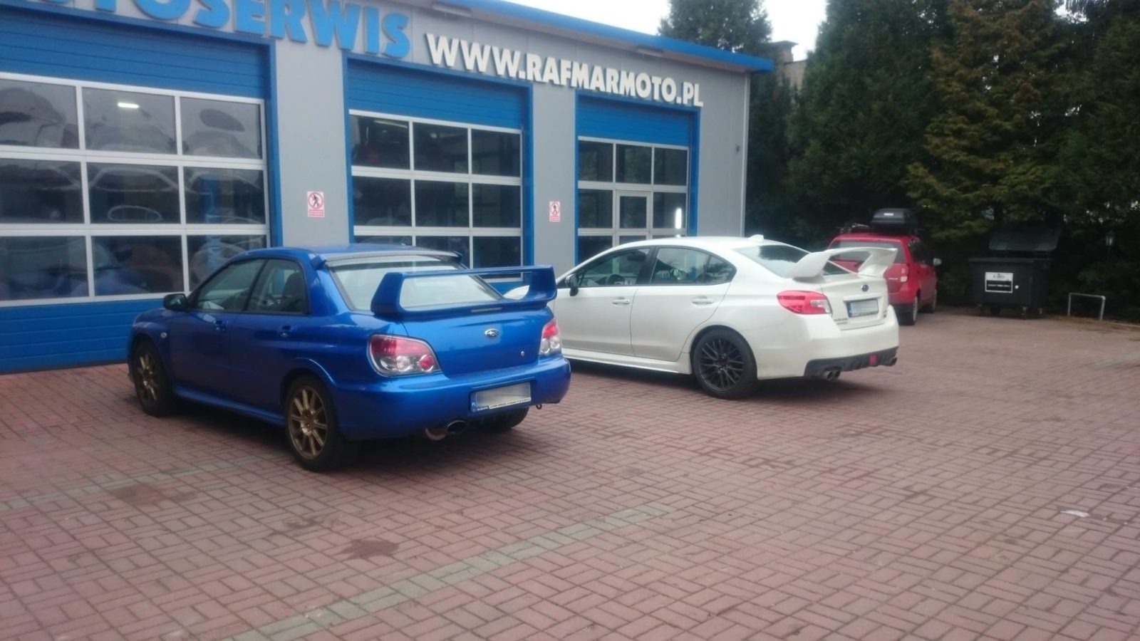 Dwa samochody Subaru, niebieski Impreza i biały WRX, zaparkowane przed warsztatem samochodowym z niebieskimi bramami i szyldem 'AUTOSERWIS WWW.RAFMARMOTO.PL'.