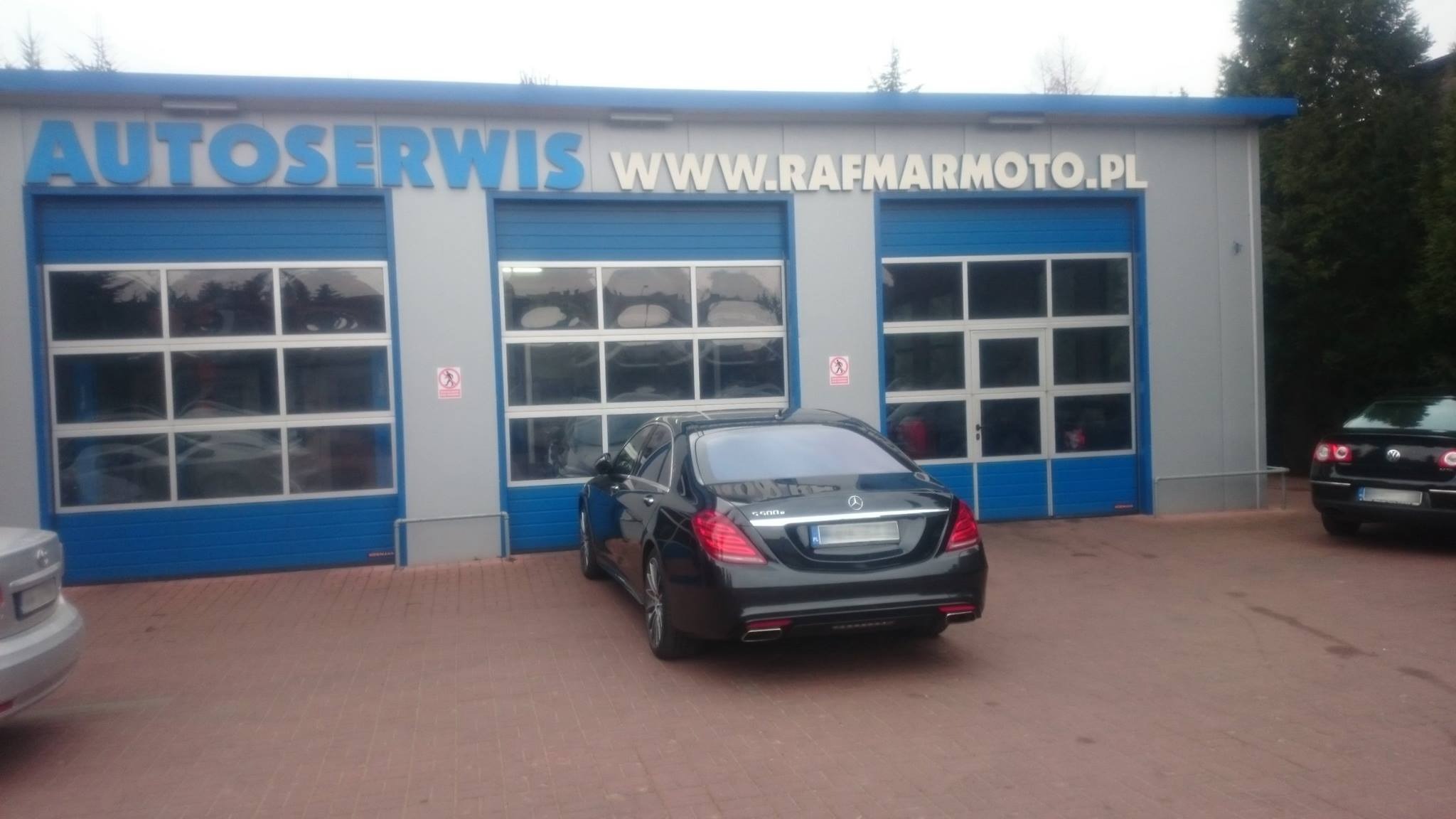 Czarny Mercedes S500 zaparkowany przed budynkiem autoserwisu z niebieskimi bramami i napisem WWW.RAFMARMOTO.PL. Widoczne inne samochody w tle.