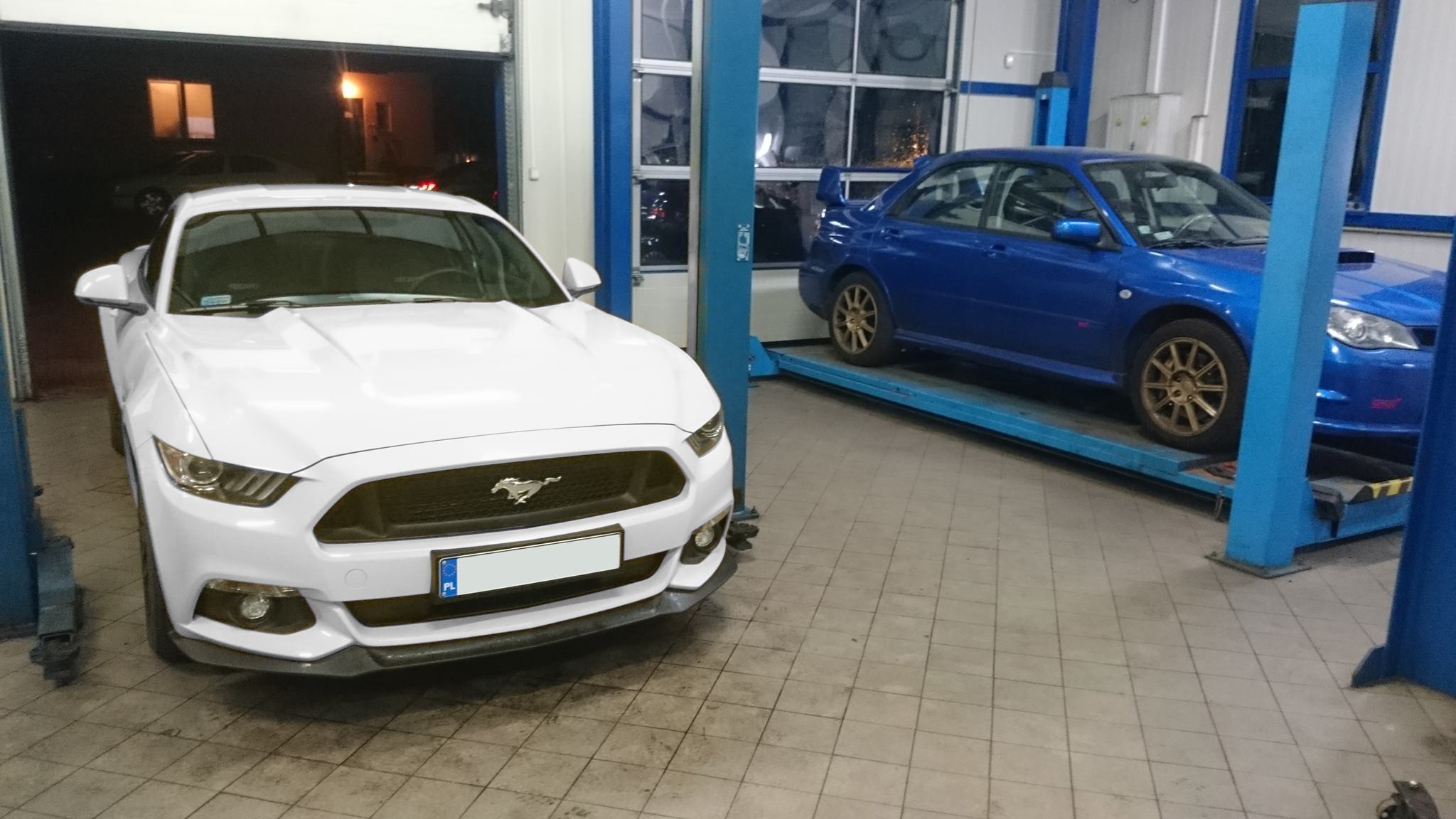 Biały Ford Mustang i niebieskie Subaru Impreza na podnośnikach w warsztacie samochodowym, widoczne narzędzia i oświetlenie warsztatowe.