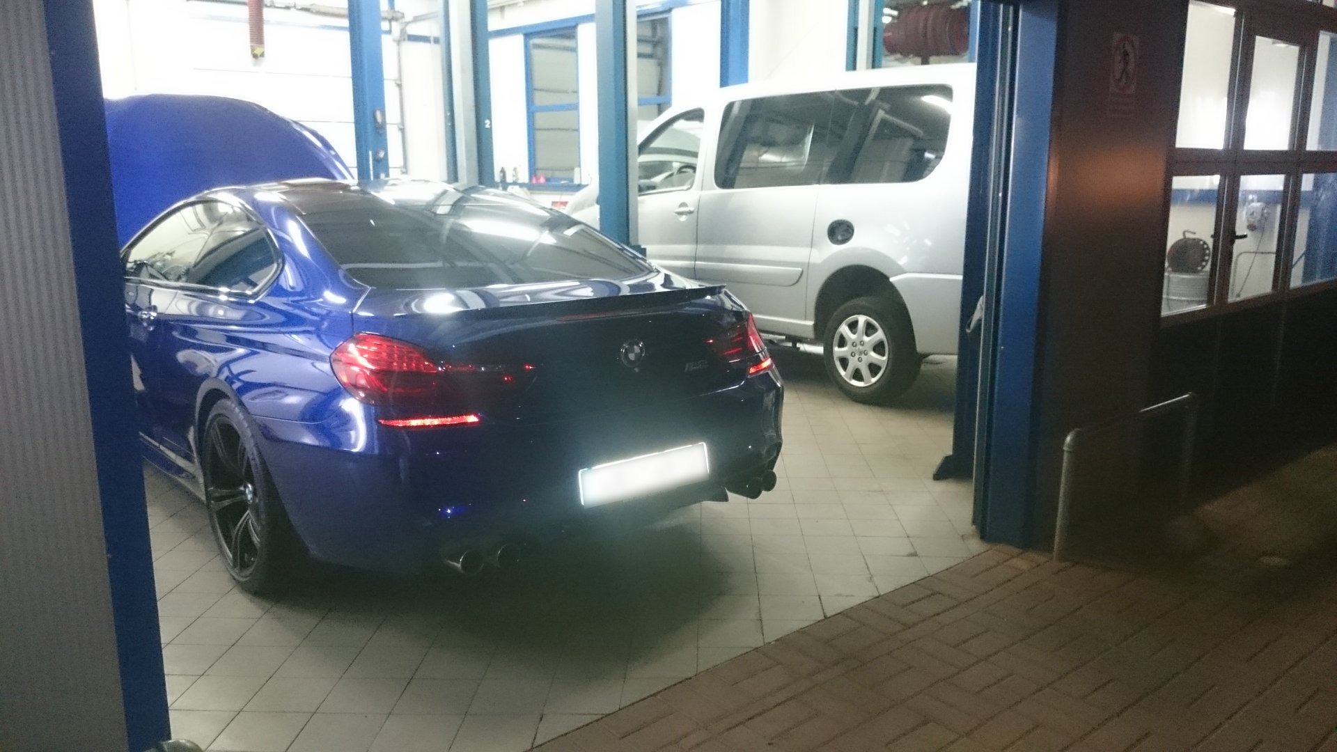 Granatowy samochód BMW z otwartą maską, stojący w warsztacie samochodowym, widoczne czarne felgi i tylne światła, w tle srebrny van.
