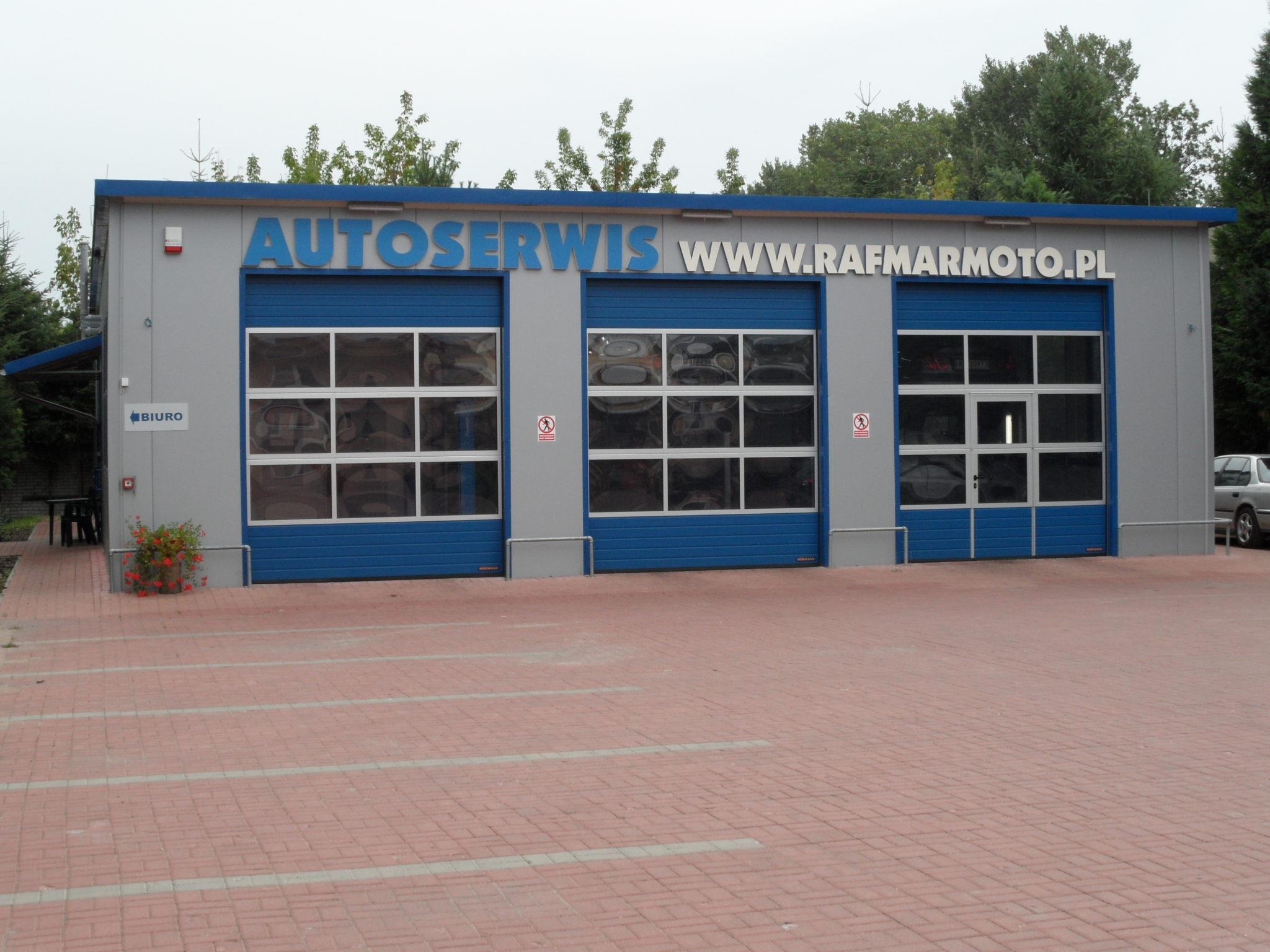 Fasada budynku autoserwisu z trzema bramami garażowymi w kolorze niebieskim i szarym, nad bramami napis AUTOSERWIS WWW.RAFMARMOTO.PL, przed budynkiem czerwona kostka brukowa