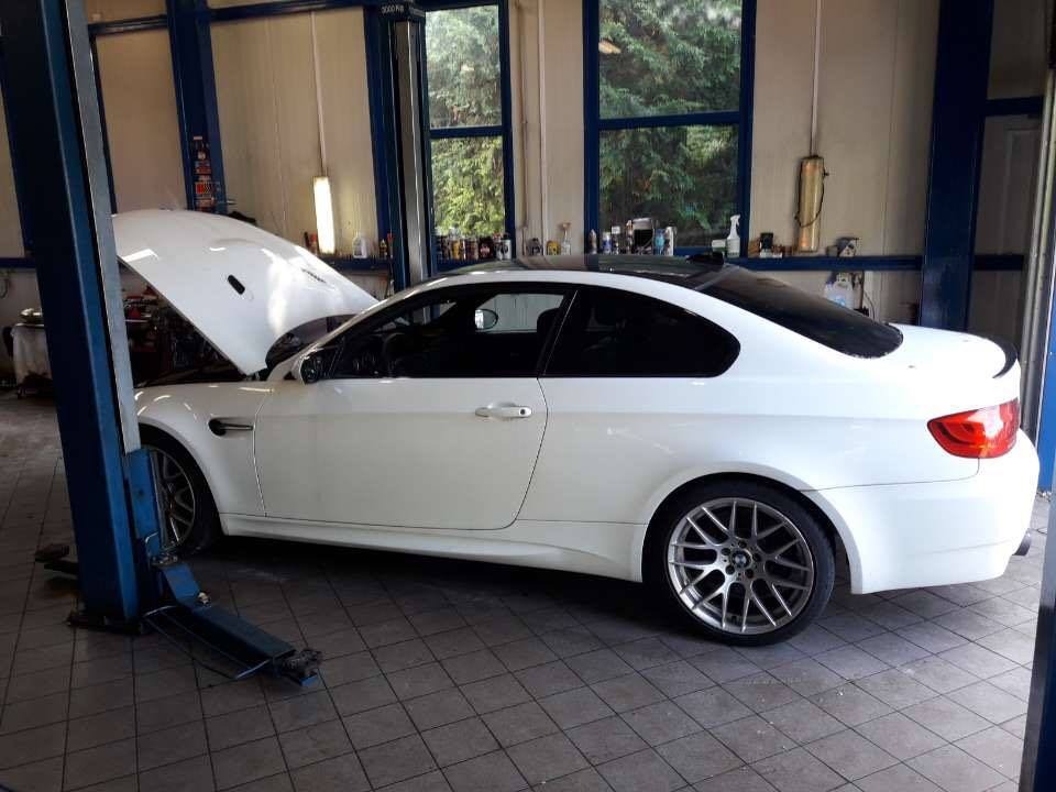 Białe sportowe auto z otwartą maską na podnośniku w warsztacie, widoczne narzędzia i części samochodowe w tle.