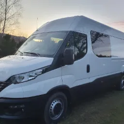 Biały bus Iveco z podwyższonym dachem na trawiastym terenie, widoczne odbicie drzew w szybach, zmierzch.