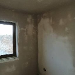 M-Plast Mateusz Zgutka - Pokój w stanie deweloperskim po nałożeniu wstępnej warstwy gładzi, widoczne niedoskonałości i fragment okna z widokiem na zewnątrz.