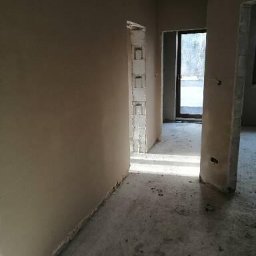 M-Plast Mateusz Zgutka - Wnętrze w trakcie prac wykończeniowych; ściany pokryte świeżym, jasnym tynkiem, widoczne surowe ościeżnice drzwiowe i betonowa posadzka.
