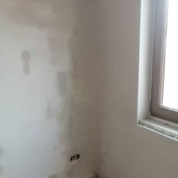 M-Plast Mateusz Zgutka - Świeżo nałożony tynk na ścianie w rogu pokoju, widoczne niedoskonałości i otwory na gniazdka elektryczne przed dalszą obróbką.