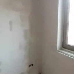 Świeżo nałożony tynk na ścianie w rogu pokoju, widoczne niedoskonałości i otwory na gniazdka elektryczne przed dalszą obróbką.