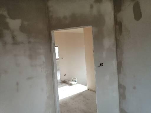 Świeżo otynkowane ściany wewnętrzne w trakcie wysychania, widoczne ślady po nakładaniu tynku, surowy stan wykończenia wnętrza z widokiem na kolejne pomieszczenie przez otwór drzwiowy.