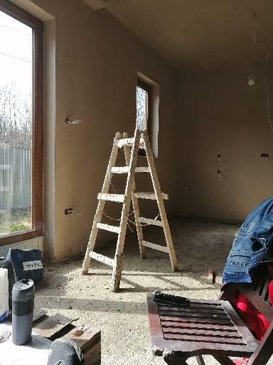 Wnętrze w trakcie tynkowania maszynowego, widoczna drabina, świeżo otynkowane ściany, wystające kable elektryczne i resztki tynku na podłodze.