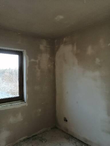 Pokój w stanie deweloperskim po nałożeniu wstępnej warstwy gładzi, widoczne niedoskonałości i fragment okna z widokiem na zewnątrz.