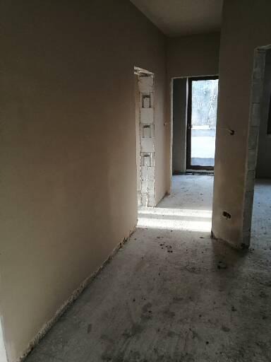 Wnętrze w trakcie prac wykończeniowych; ściany pokryte świeżym, jasnym tynkiem, widoczne surowe ościeżnice drzwiowe i betonowa posadzka.