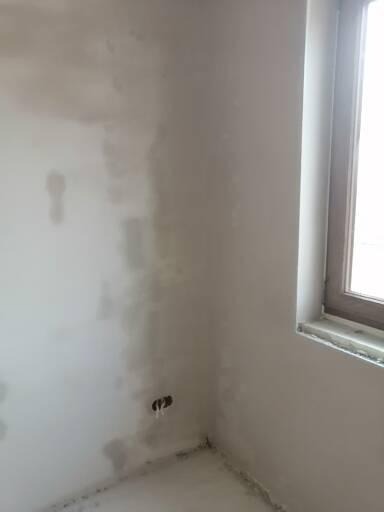 Świeżo nałożony tynk na ścianie w rogu pokoju, widoczne niedoskonałości i otwory na gniazdka elektryczne przed dalszą obróbką.