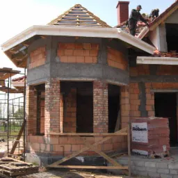 Budowa domu w stanie surowym otwartym: murowane ściany z czerwonej cegły, widoczne rusztowania, pracownicy na dachu, materiały budowlane Roben na palecie.