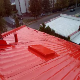 Czerwony, metalowy dach z kominem i wyłazem dachowym, widok z góry na tle zabudowy miejskiej.