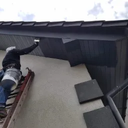 Pracownik na drabinie maluje podbitkę dachu na ciemny kolor, widoczny kubeł z farbą i zabezpieczenia.