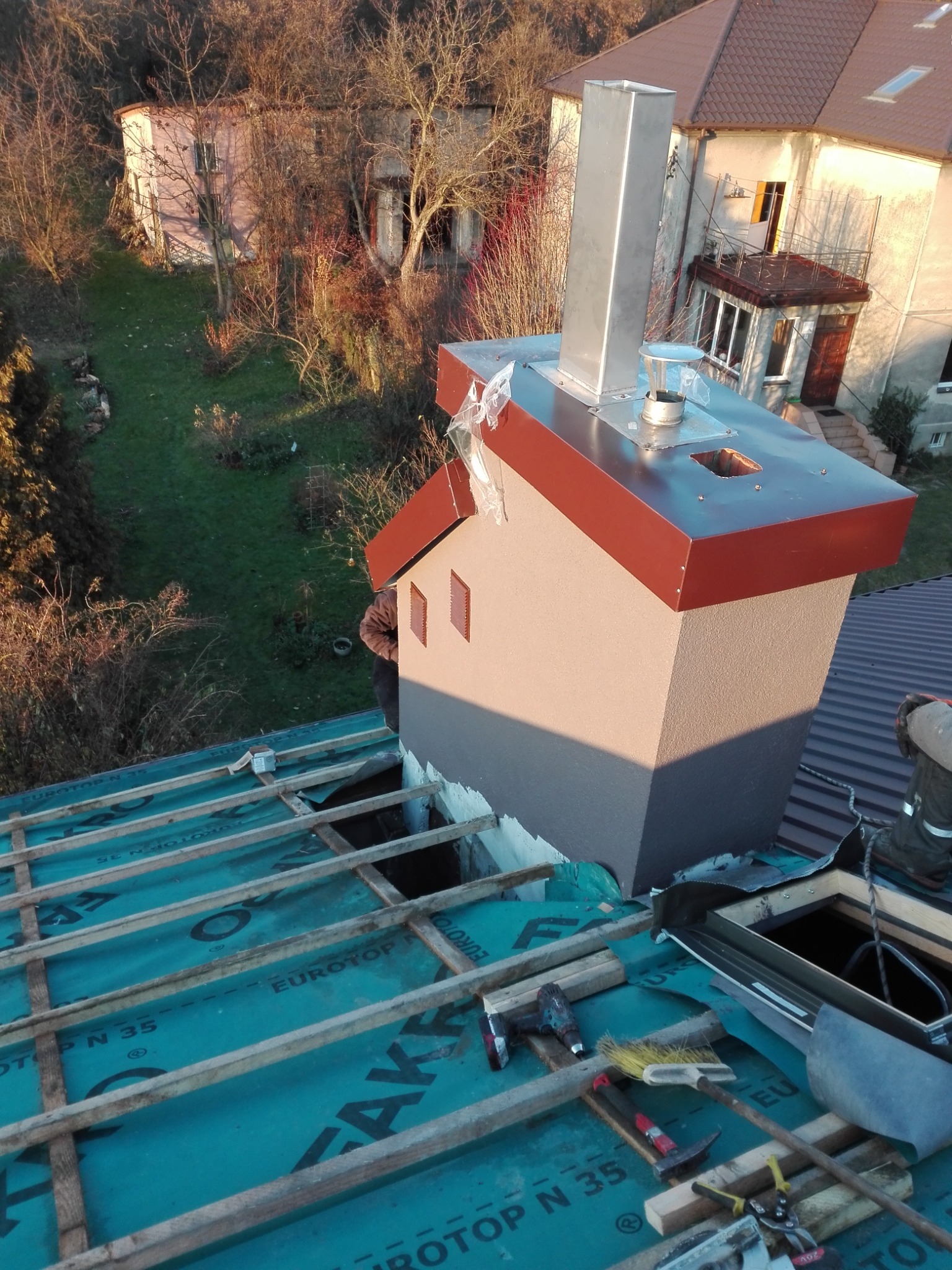 Montaż komina z metalowym wkładem na dachu z membraną, widoczne narzędzia i fragment sylwetki pracownika, w tle budynki mieszkalne i drzewa.