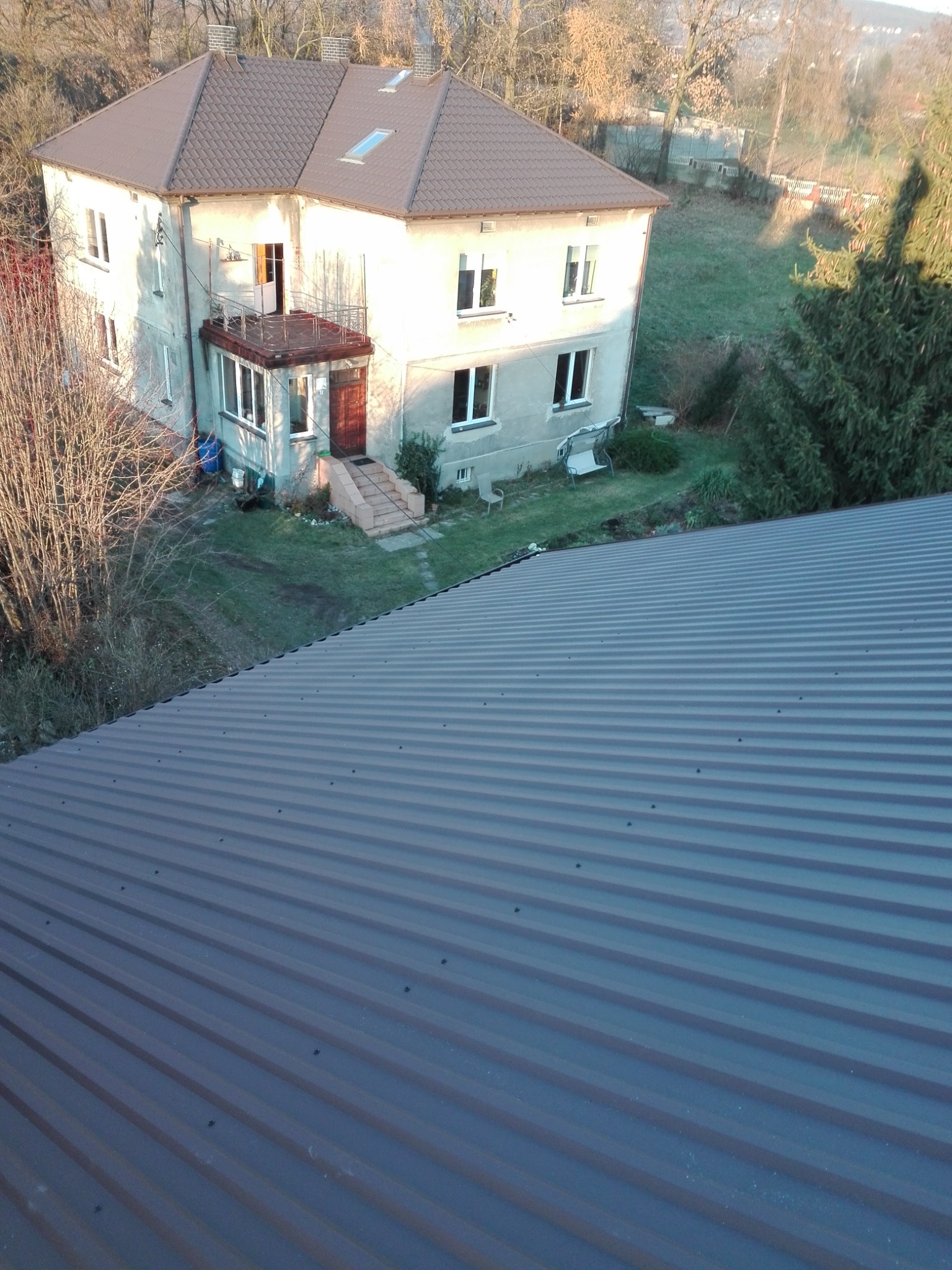 Widok z góry na nową, brązową blachodachówkę na dachu domu jednorodzinnego z ogrodem, w tle drzewa i budynki.