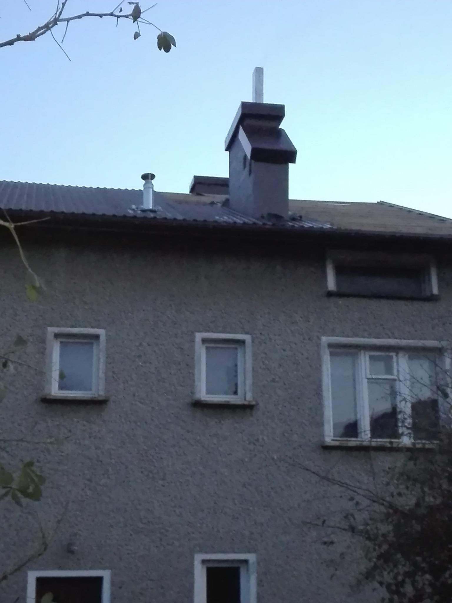 Szary dom z oknami, widoczny dach z kominem i rurami wentylacyjnymi, ujęcie z dołu.