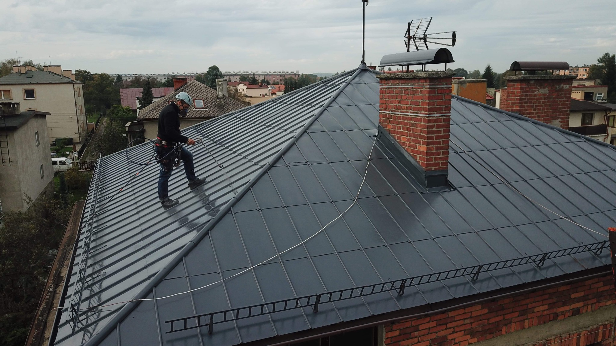 Pracownik w uprzęży zabezpieczającej, stojący na nowym, grafitowym dachu z blachy trapezowej, w trakcie prac na wysokości, z widocznym kominem z czerwonej cegły i anteną telewizyjną.