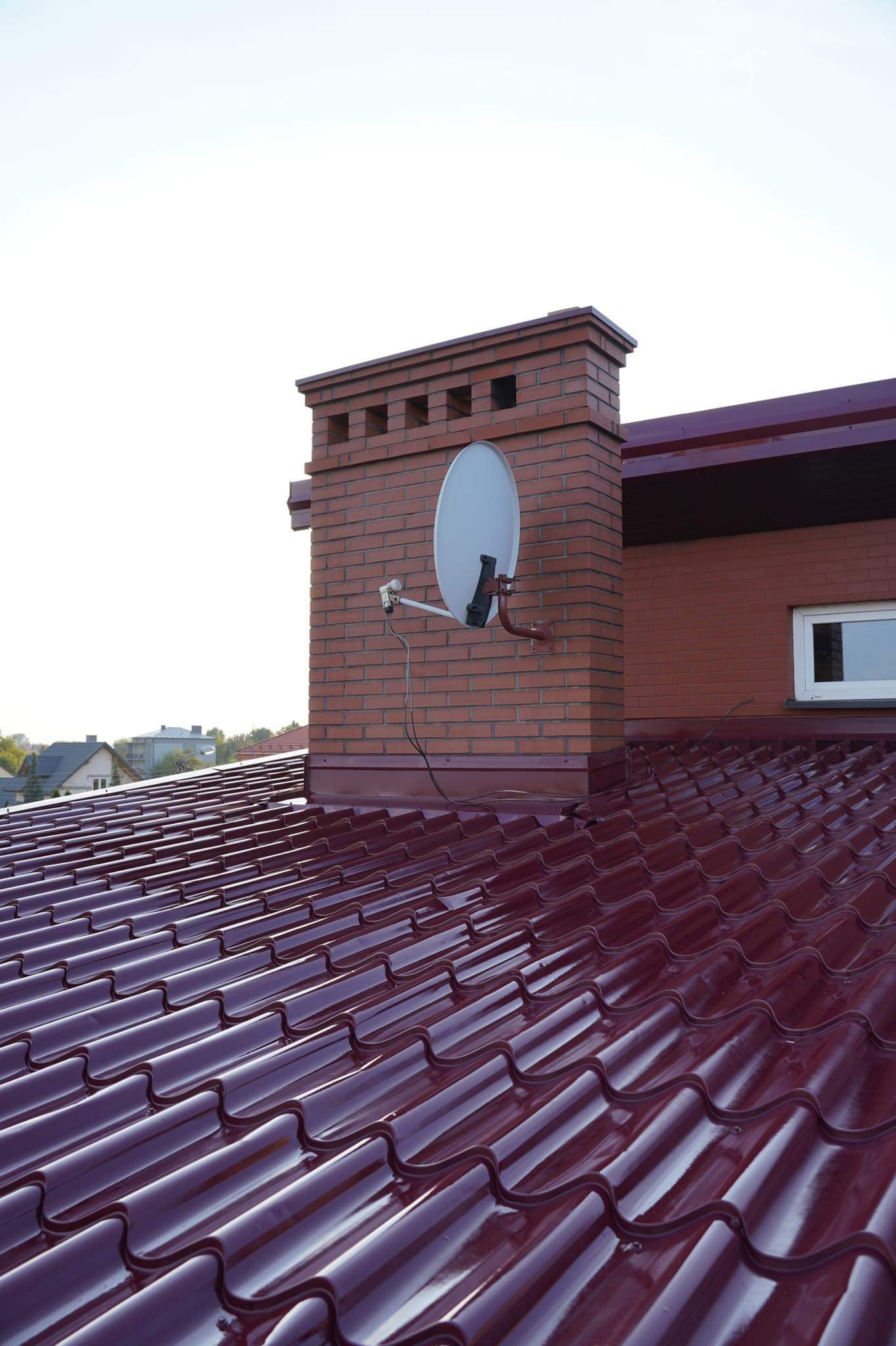 Czerwona, metalowa dachówka na dachu domu z ceglanym kominem i anteną satelitarną.