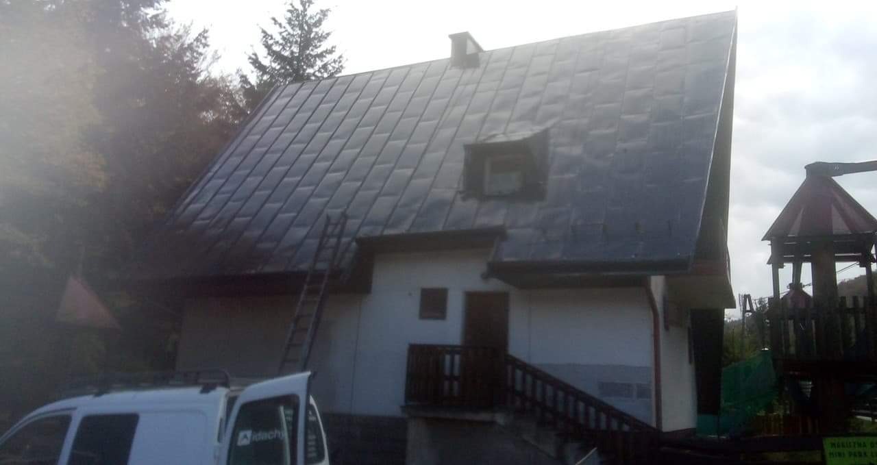 Dom z blachy trapezowej na dachu, drabina oparta o ścianę, częściowo widoczny samochód dostawczy z logo firmy dekarskiej, plac zabaw w tle.