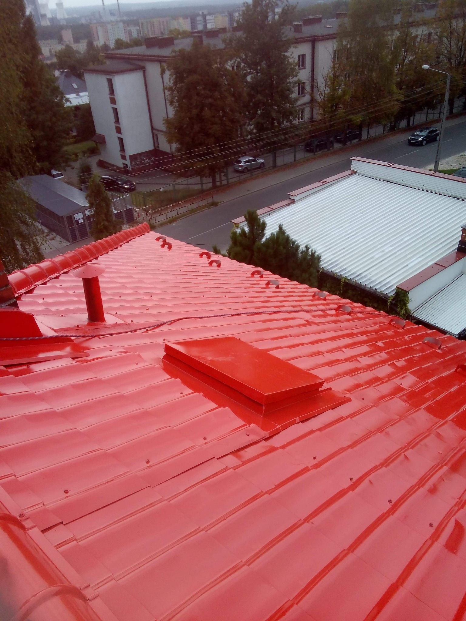 Czerwony, metalowy dach z kominem i wyłazem dachowym, widok z góry na tle zabudowy miejskiej.