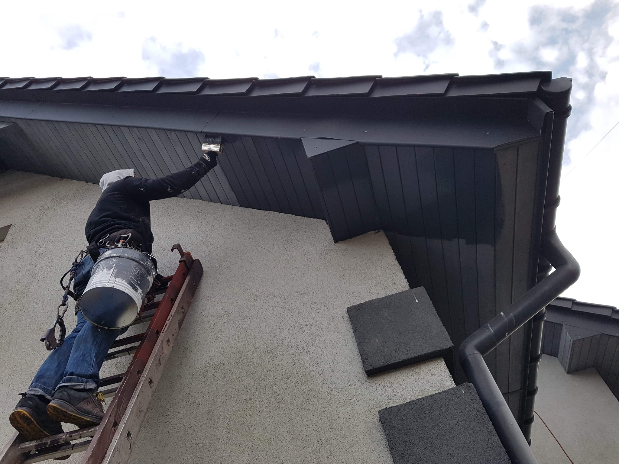 Pracownik na drabinie maluje podbitkę dachu na ciemny kolor, widoczny kubeł z farbą i zabezpieczenia.