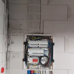 Rozdzielnica elektryczna z licznymi przewodami ułożonymi w peszlach i listwach na surowej ścianie z betonu komórkowego.